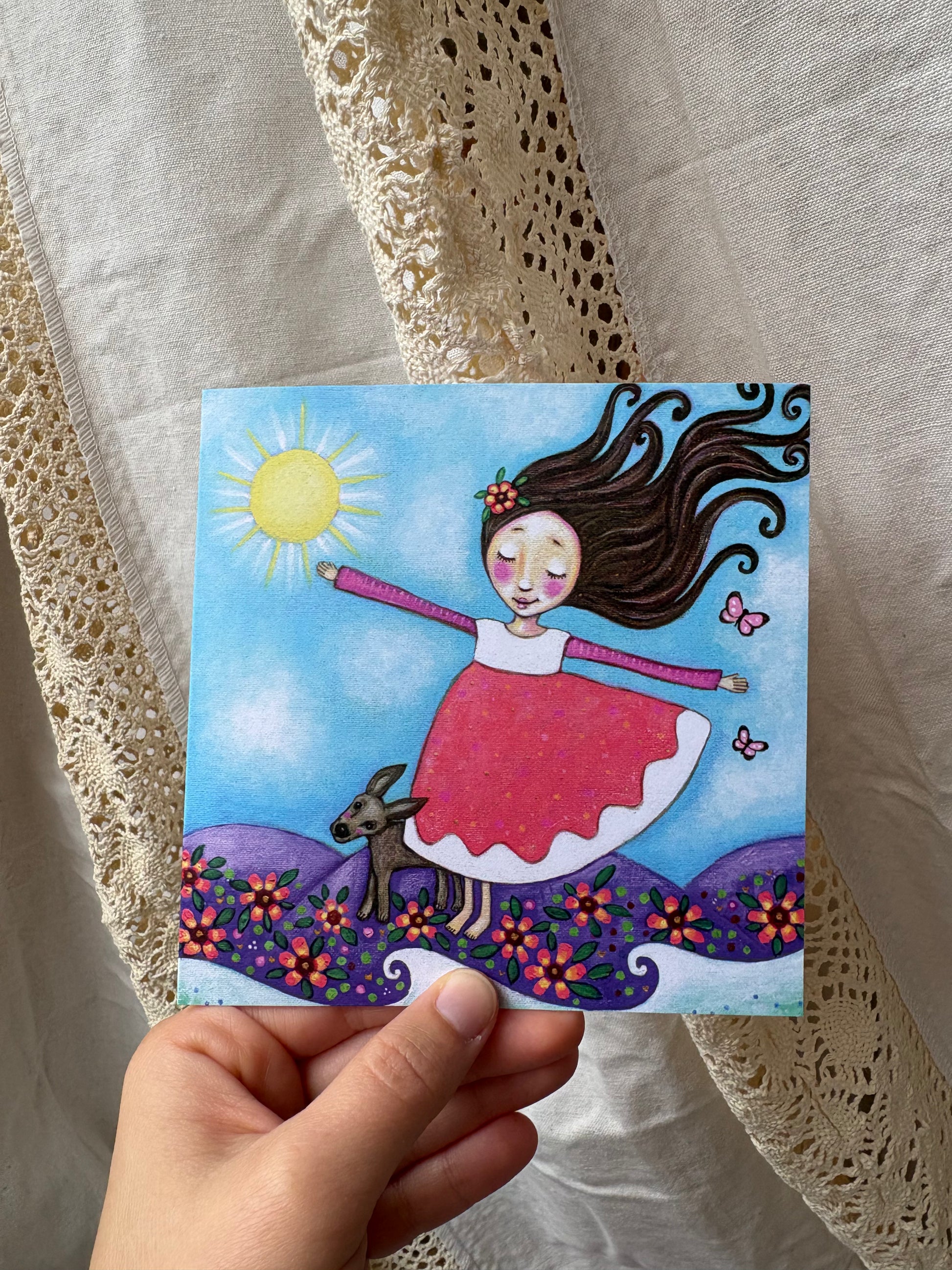 Freedom greeting card: hand-painted illustration of a girl standing with her eyes closed facing toward the sun with the wind blowing in her hair, a blue sky, butterflies flying beside her, flowers surrounding her feet, the ocean below her and a brown dog standing next to her. 