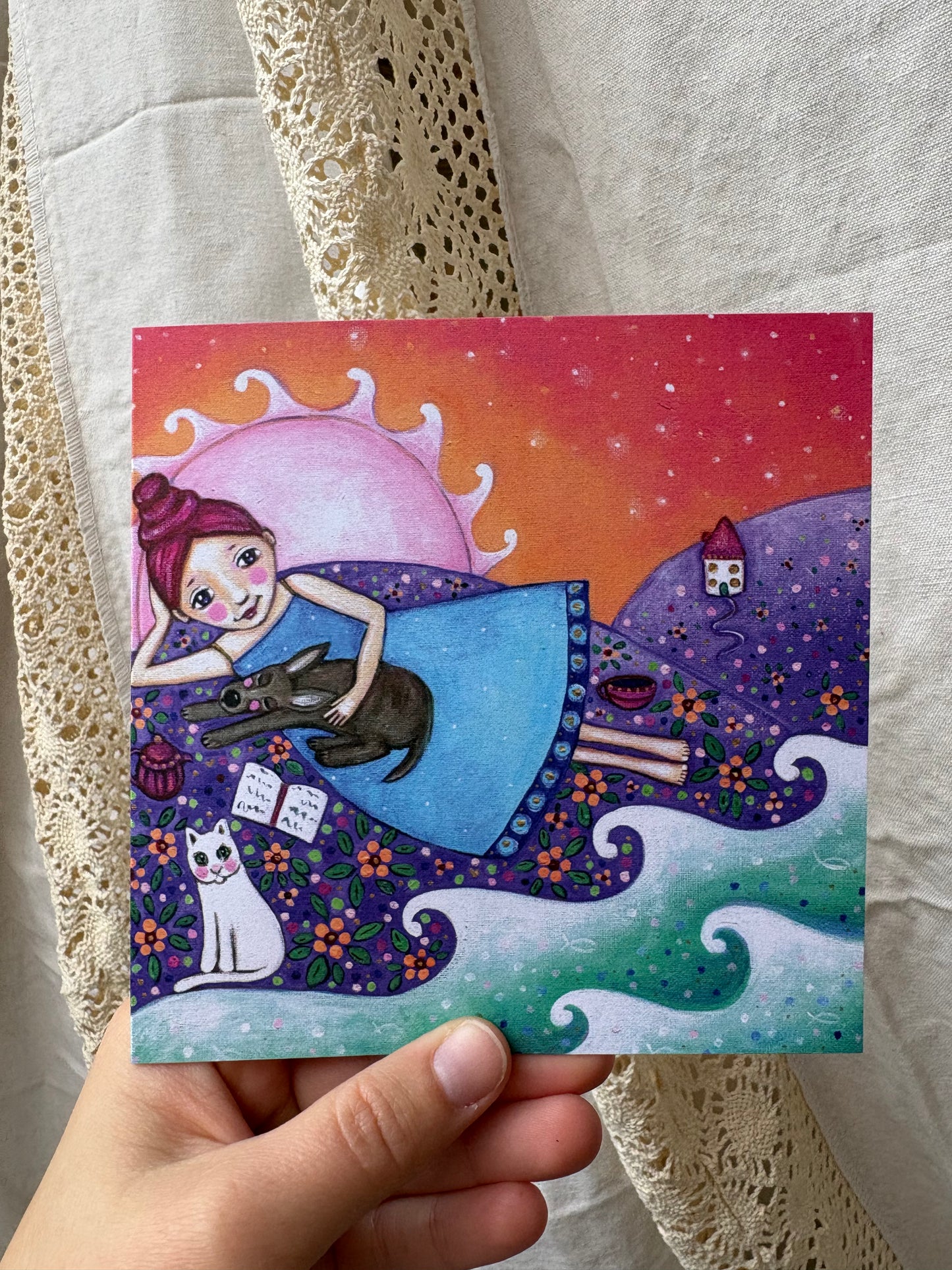 Home greeting card: hand-painted illustration of a woman lying on a hill with a pot of tea and an open book petting a sleeping dog, with a white cat, a distant cottage on the hill and ocean waves, with the setting sun behind her, from What Makes Your Heart Sing